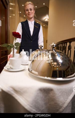 Lächelnde junge Frau auf dem Wagen, die warmes Essen den Hotelflur hinunterträgt Stockfoto