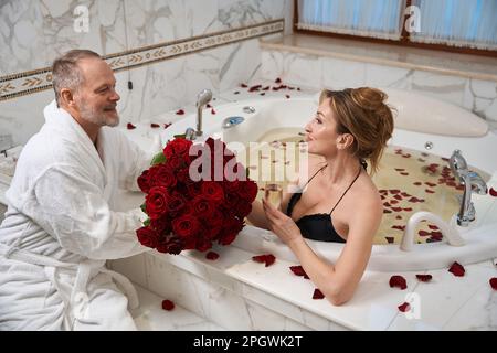 Ein lächelnder Mann im Bademantel gibt seiner reizenden Frau einen Blumenstrauß Stockfoto