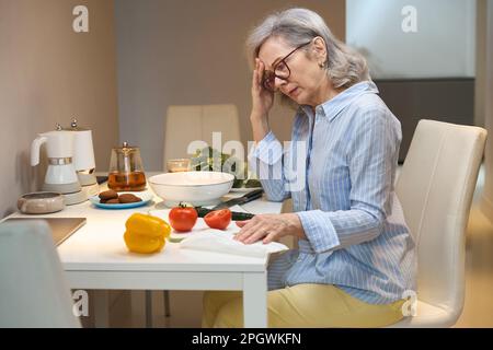 Traurige grauhaarige Dame in lässiger Kleidung sitzt auf dem Akitchen Tisch Stockfoto