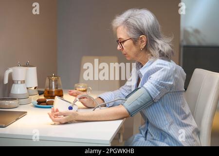 Die grauhaarige Dame sitzt am Küchentisch und misst ihren Blutdruck Stockfoto
