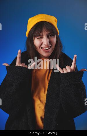 Vertikales Studioporträt einer jungen Frau in einem gelben Hemd, einer schwarzen Jacke und einer gelben Kappe, die mit beiden Händen, blinzelnd und hängend eine „Liebesgeste“ macht Stockfoto