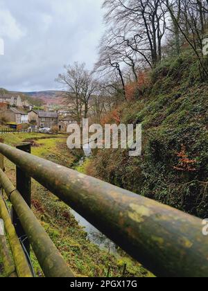 Eine ländliche Szene mit einem Holzzaun im Vordergrund, einem Bach, der daneben verläuft, und fernen Gebäuden im Hintergrund Stockfoto