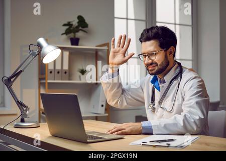 Porträt eines glücklichen, lächelnden Arztes, der während einer Online-Konferenz an seinem Arbeitsplatz sitzt und per Webcam-Videoanruf auf einem Laptop telefoniert. Stockfoto