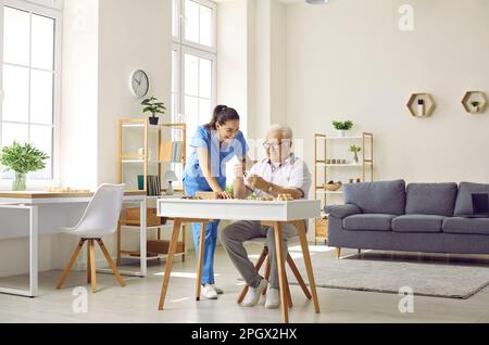 Krankenschwester im Seniorenheim hilft Seniorenmann, der am Schreibtisch sitzt und Puzzles macht Stockfoto