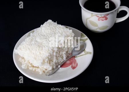 Ein Stück Couscous in einer Porzellan-Untertasse auf schwarzem Hintergrund, typisch brasilianisches Gericht mit Tapioka, Milch, Zucker und Kokosmilch. Mit einer Tasse Kaffee Stockfoto