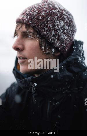 Porträt eines jungen Mannes mit Wollbeere und schwarzer Jacke unter den Schneeflocken im Winter. Stockfoto