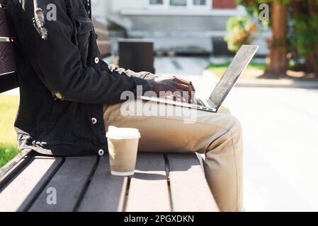 Arbeiten mit einem Laptop. Nahaufnahme eines afroamerikanischen Mannes in schwarzer Jacke im Freien in der Stadt, der auf einer Bank sitzt Stockfoto