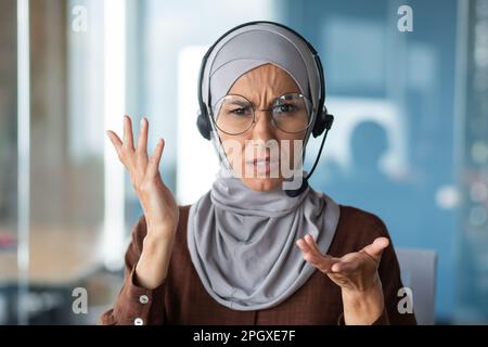 Verärgerte Frau im Hijab, die bei Videogesprächen spricht, Geschäftsfrau mit Headset-Telefon, die aus nächster Nähe auf die Webkamera schaut und mit Kollegen in einem Online-Meeting spricht. Stockfoto