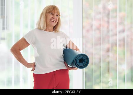 Positive Seniorin in Sportkleidung im Innenbereich im Stehen und Halten der Matte in den Händen Stockfoto