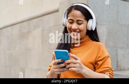 Glückliche Frau aus Südostasien, die im Stadtzentrum mit ihrem Smartphone und ihren Kopfhörern Musik hört Stockfoto