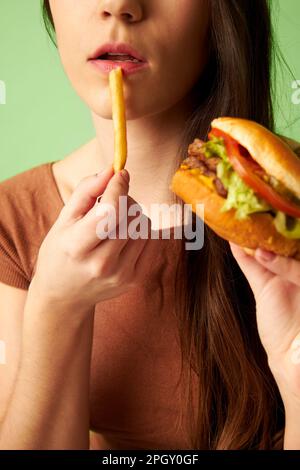 Porträt einer Frau, die einen Hamburger-Cheeseburger hält, während sie die untere Hälfte des Gesichts isst Stockfoto