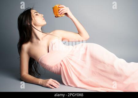 Porträt einer Frau, die einen Hamburger hält und Haare im Wind bläst, während sie sich im Ballkleid legt Stockfoto