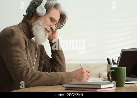 Mann mittleren Alters mit Laptop und Kopfhörern, der drinnen am Tisch lernt Stockfoto
