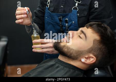 Friseur mit Bartöl in der Nähe des Kunden im Friseursalon, Schließung. Professioneller Rasierservice Stockfoto