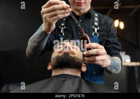 Friseur mit Bartöl in der Nähe des Kunden im Friseursalon, Schließung. Professioneller Rasierservice Stockfoto