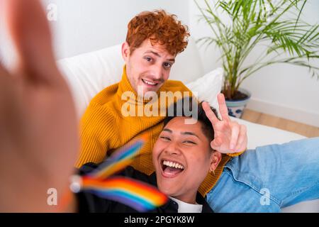 Wunderschönes schwules Paar, das zu Hause romantisch ist, auf dem Sofa, schwules Paar, das Selfie macht und Siegeszeichen macht, lgbt-Konzept Stockfoto