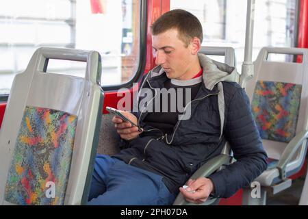 Der junge Kerl sitzt im Auto mit dem Telefon in der Hand und sieht sich das Video online an Stockfoto