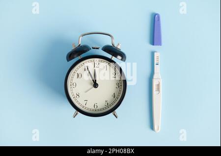 Stillleben mit Wecker und Schwangerschaftstest am Arbeitsplatz - zwei Streifen auf blauem Hintergrund. Endlich schwanger Stockfoto