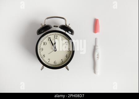 Stillleben mit schwarzem Vintage-Wecker und Inkjet-positivem Schwangerschaftstest auf isoliertem weißem Hintergrund Stockfoto