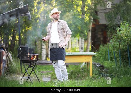 Grill Zeit. Europäischen Mann in hat Warten auf Gegrilltes. Er sitzt in der Nähe von Feuer und anderen. Stockfoto