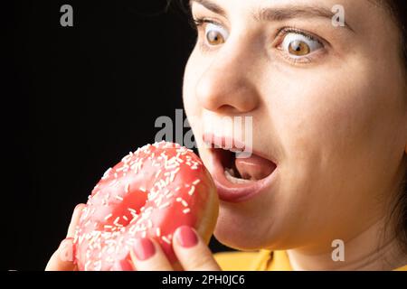 Eine Frau beißt einen großen roten Donut, einen schwarzen Hintergrund, ein Ort für Text. Gluttonie, überfressen und zuckersüchtig Stockfoto