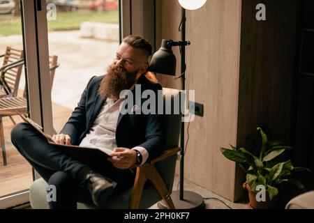 Ein gutaussehender bärtiger Mann, der etwas auf seinen Notizblock schreibt, mit überkreuzten Beinen, der bequem auf dem Stuhl im Café sitzt Stockfoto