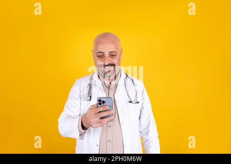 Der Hausarzt hält das Telefon in der Hand. Ein Typ mittleren Alters, der Technologie benutzt. Positive lächelnde Arztbotschaft. Mit der Gesundheitsklinik Stockfoto