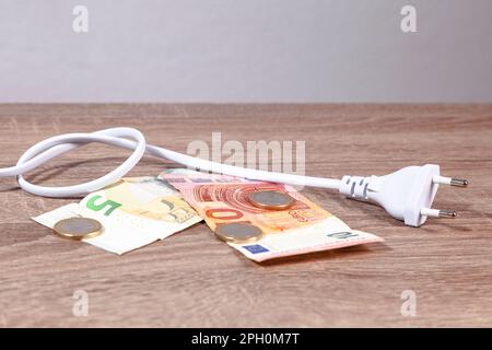 Weißes Stromkabel mit europäischem Stecker und Euro-Banknoten und -Münzen über einem Holztisch vor einer weißen Wand. Stockfoto