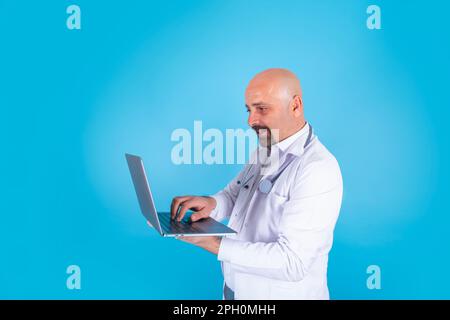 Das Porträt des Kaukasischen Arztes. Stehender Arzt hält und tippt Laptop, isolierter blauer Hintergrund, Kopierraum. Er trägt einen weißen Mantel Stockfoto