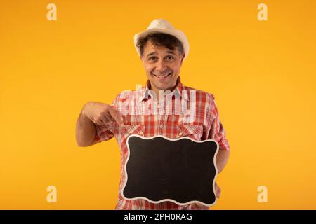 Schwarzhaariger Mann mit Hut und farbenfrohem Hemd, lächelnd, mit einem Black-Board-Schild in den Händen Stockfoto