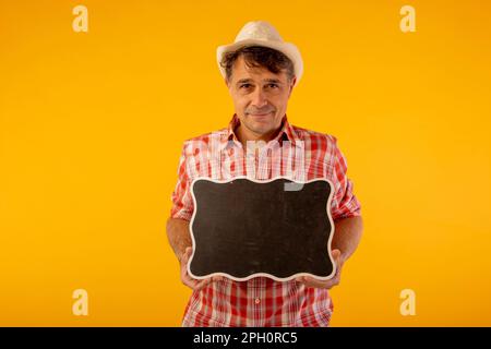 Schwarzhaariger Mann mit Hut und farbenfrohem Hemd, lächelnd, mit einem Black-Board-Schild in den Händen Stockfoto