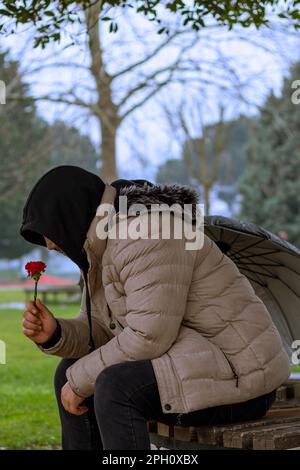 Ein Mann mit Schirm sitzt unter dem Baum, hält rote Blume auf der Bank, regnerisch Stockfoto