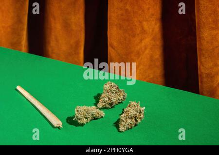 Drei trockene Knospen medizinisches Marihuana liegen auf dem grünen Tisch neben dem Gelenk. Vor dem Hintergrund brauner Samtvorhänge Stockfoto