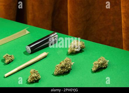 King-Size-Joint, Papier und Feuerzeug liegen zwischen trockenen Marihuana-Knospen auf einem grünen Tisch vor einem Hintergrund mit braunen Samtvorhängen Stockfoto