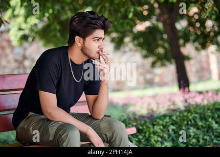 Junger indischer Raucher Porträt in schwarzem T-Shirt und silberner Halskette auf einer Bank im öffentlichen Park, hinduistischer rauchender Mann Porträt. Hübscher indianer Stockfoto