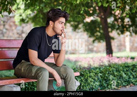 Junger indischer Raucher Porträt in schwarzem T-Shirt und silberner Halskette auf einer Bank im öffentlichen Park, hinduistischer rauchender Mann Porträt. Hübscher indianer Stockfoto