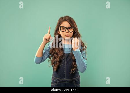 Ein seriöses Teenager-Mädchen, inspiriert von Idee, hat lockiges Haar halten Party Accessoire Brille, Party. Stockfoto