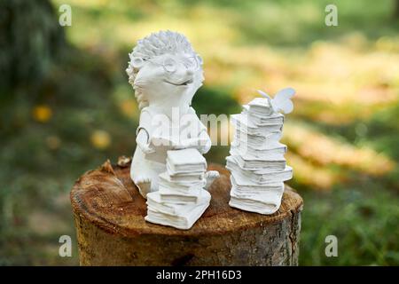 Die kleine Igelstatue neben dem Bücherstapel befindet sich auf einem Holzstumpf auf grünem Hintergrund, süße Kinder machen Igelfiguren über Lernen und Bücher readi Stockfoto