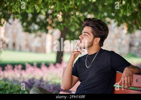 Junger indischer Raucher Porträt in schwarzem T-Shirt und silberner Halskette auf einer Bank im öffentlichen Park, hinduistischer rauchender Mann Porträt. Hübscher indianer Stockfoto