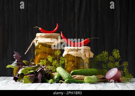 Eingelegte Gurken in einem Glas, frische Gurken, rotes und grünes Chili, Dill, Basilikum, Knoblauch auf dunklem Holzhintergrund. Paprika, Zwiebeln und frische Cornflakes Stockfoto