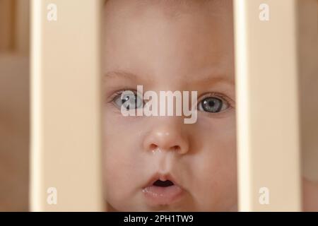 Nahaufnahme eines weinenden süßen Babys im Kinderbett zu Hause. Ein kleines Mädchen mit Tränen in den Augen steht in seinem Kinderbett. Schutz der Rechte des Kindes. Kind-fa Stockfoto