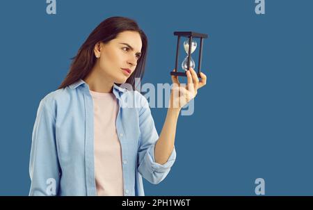 Wunderschöne junge braune Frau, gekleidet in einem hellen T-Shirt und einem hellblauen, übergroßen Hemd, starrt aufmerksam auf die Sanduhr in ihrer Hand. Stockfoto