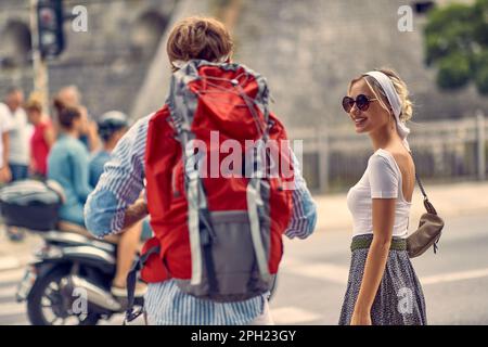 Eine hübsche blonde Frau, die einen Mann mit Rucksack ansieht. Sommerspaziergang in der Stadt. Touristen, Paare, Reisen, lustiges Konzept. Stockfoto