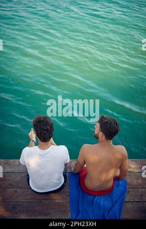 Zwei junge Männer sitzen auf einem Holzsteg und schauen auf das Wasser. Hochwinkelschuss. Urlaub, Spaß, Zusammensein, Lifestyle-Konzept. Stockfoto