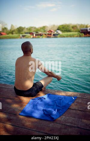 Ein Mann, der morgens auf dem Wasser meditiert. Junger Mann, der auf einem hölzernen Steg am Meer sitzt und spirituelle Übungen macht. Entspannung, Disziplin, Lifestyle-Konzept. Stockfoto