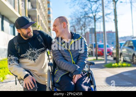 Ein Begleiter, der fröhlich mit einem jungen Mann im Rollstuhl auf dem Bürgersteig spricht Stockfoto