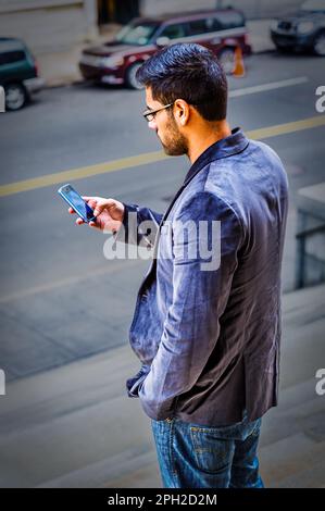 Ein junger Gelehrter mit einem kleinen Bart und Schnurrbart steht auf der Straße und überprüft mit einer Brille und einer Hand die Nachrichten auf einem Stockfoto
