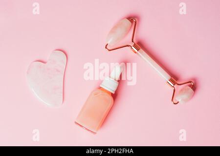 Gesichtswalze aus Rosenquarzkristall und Gua-Sha-Schaber, Gesichtsserum oder organisches Kosmetiköl auf rosa Hintergrund. Gesichtsmassageset für die Lifttherapie. Stockfoto