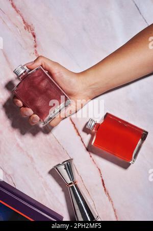 Eine Frauenhand hat zwei Glasgefäße mit Rum-Cocktails, Gin, kosmopolitischer Stil, hartes Licht auf einem elfenbeinfarbenen Tisch mit Barzubehör, eine Hand Stockfoto