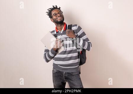 Stolzer afrikanischer Student mit Brille, gestreiftem Pullover und Jeans, mit Rucksack und Notizbüchern, mit kurzen geflochtenen Haaren, die gegen einen li stehen Stockfoto